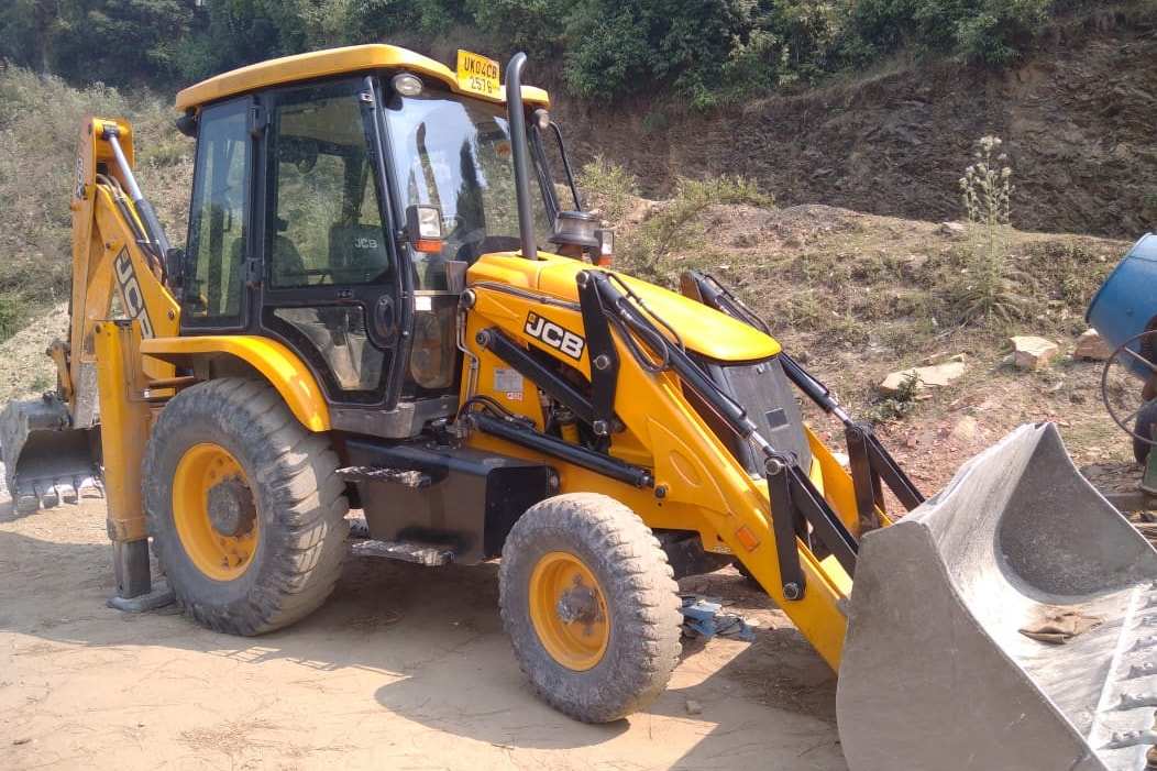 Front three-quarter view of RBE’s JCB backhoe loader parked below a cut hill slope on a hillside site at Padampuri, Nainital district.
