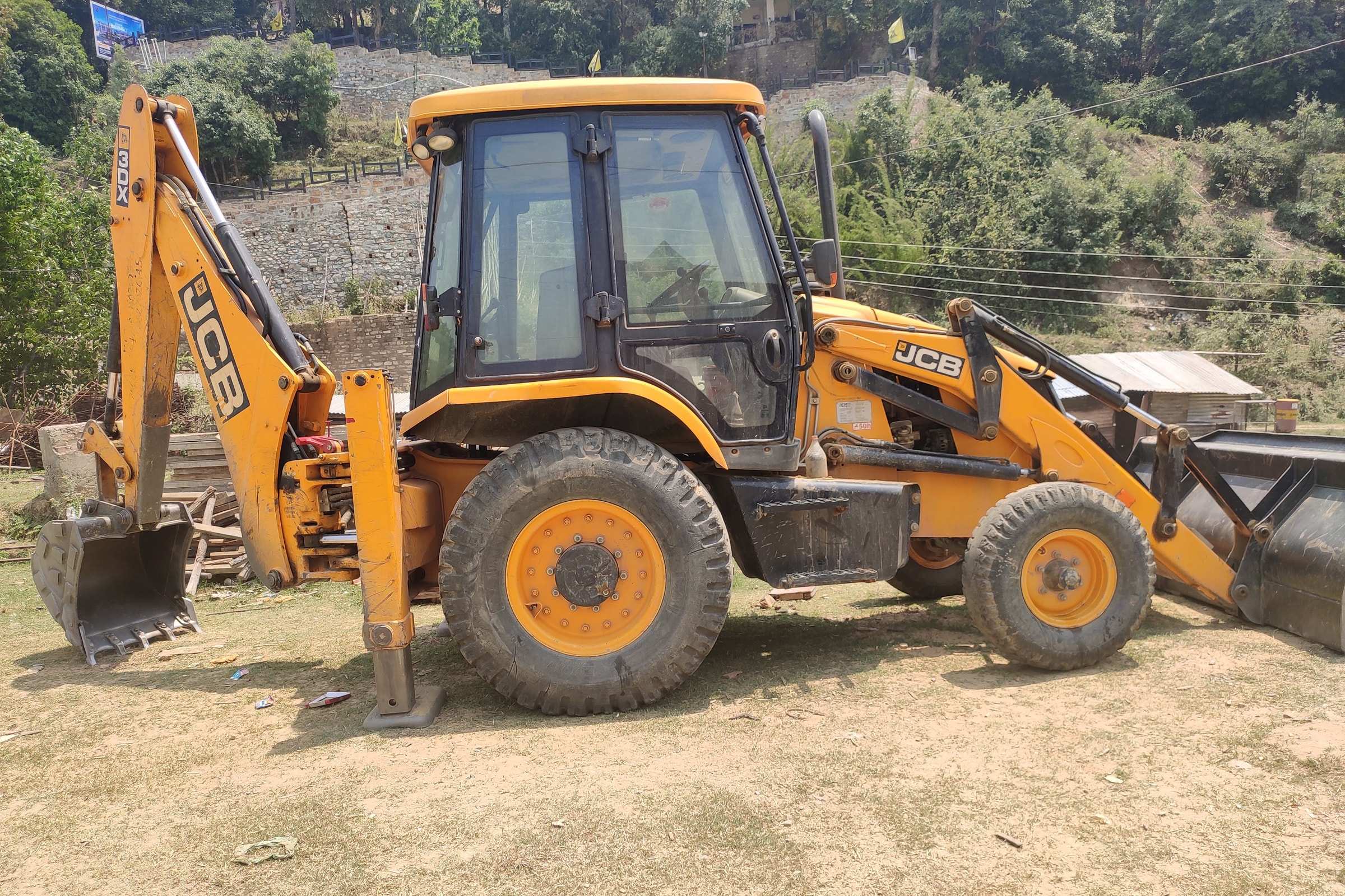 JCB backhoe loader with stabilisers planted on firm ground below a stone retaining wall on a hillside site at Padampuri.