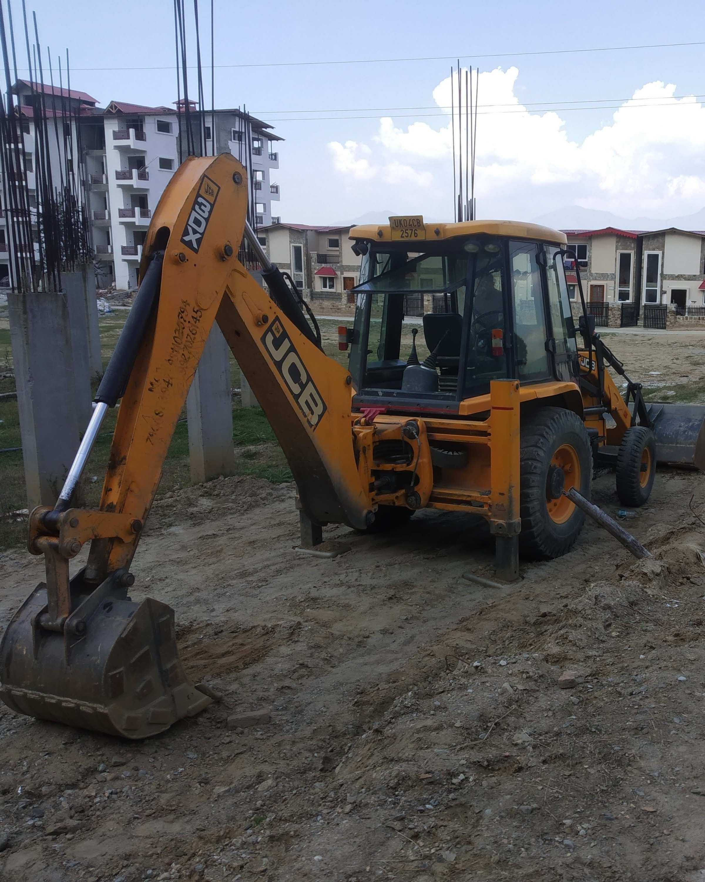 Rear view of a JCB backhoe loader with stabilisers planted on a building construction site, rebar columns and apartment blocks behind, Padampuri.