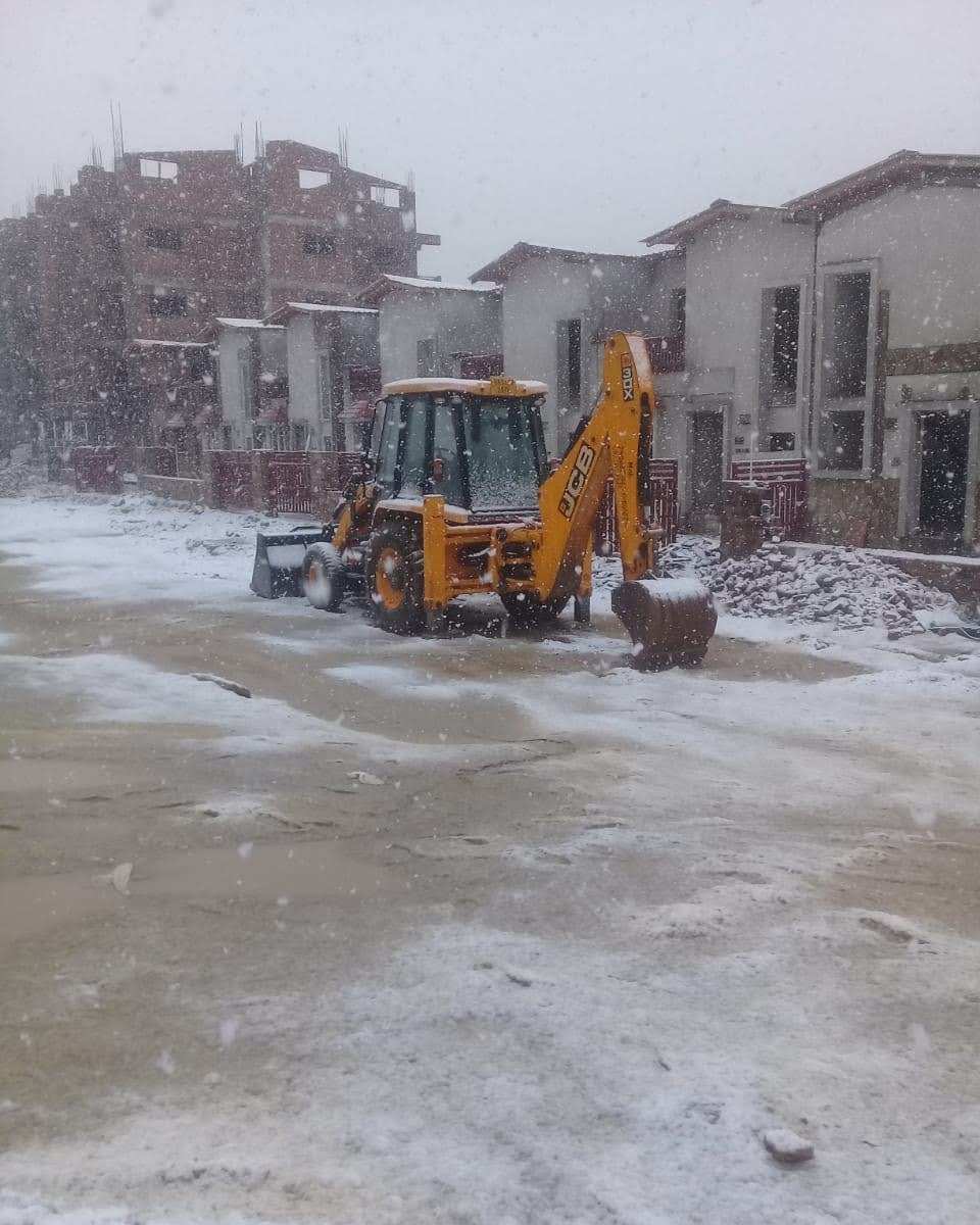 JCB backhoe loader working through snowfall on the Ranikhet–Majkhali resort construction site, with finished cottages and a building under construction behind.